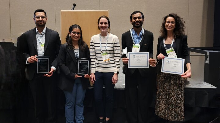 Five award recipients and a presenter pose on a stage at a national entomology conference. From left to right: Sajjan Grover and Kashish Verma hold plaques, Anjel Helms stands in the center as the awards presenter, and Sanket Shinde and Andrea Rilakovic hold certificates.